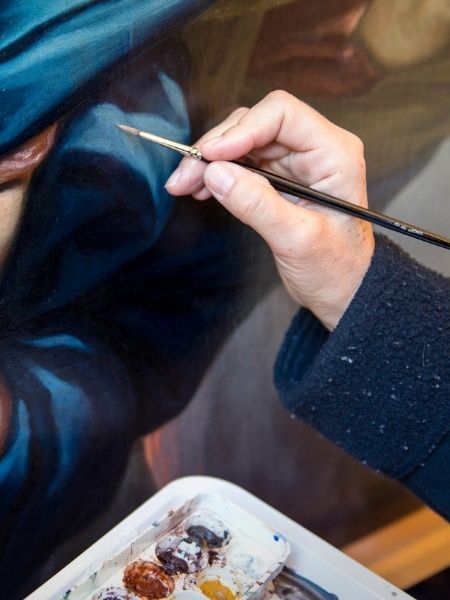 Close-up of a conservator's gloved hand using a fine brush to restore a detail on an oil painting