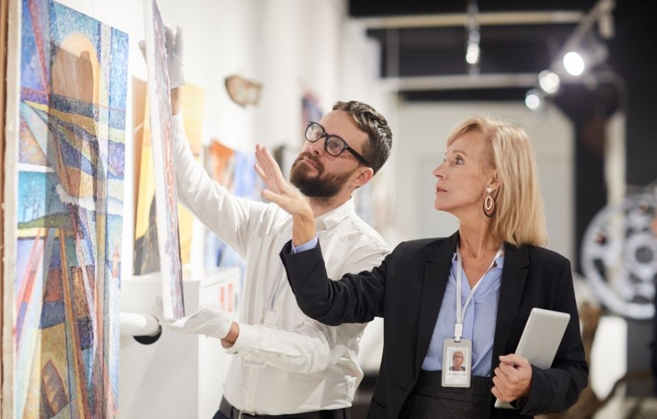 Two collections staff members examining a painting in a gallery, with one wearing gloves and holding a tablet to support conservation documentation