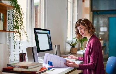 A collections staff member reviewing printed documents at a desk with a desktop computer, illustrating day-to-day cataloguing and inventory management workflows