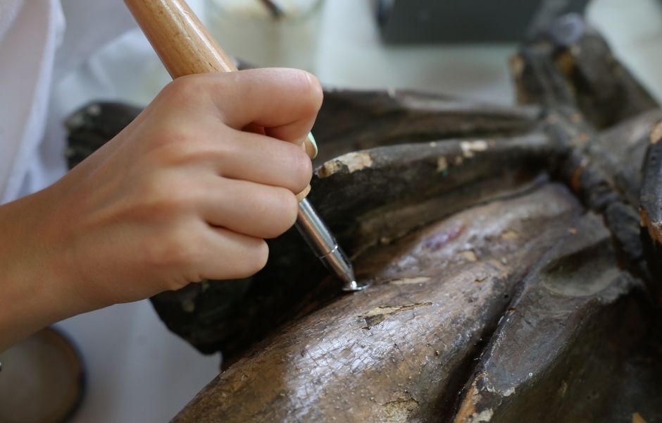 A conservator in a white coat applying treatment to a damaged wooden sculpture, representing the hands-on conservation work documented within TMS Conservation Studio