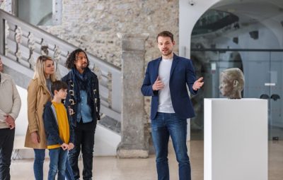 A guide presenting a sculpted bust on a plinth to a small group of visitors in a stone-walled gallery space