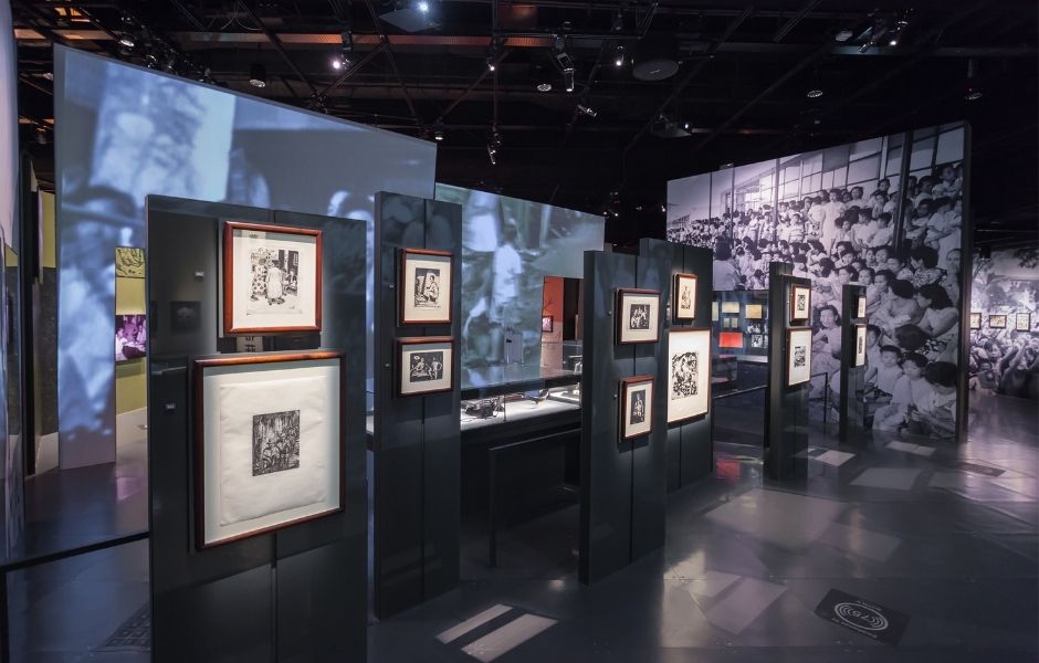 A dramatic exhibition space displaying framed prints on dark panels with large-scale photographic projections covering the surrounding walls