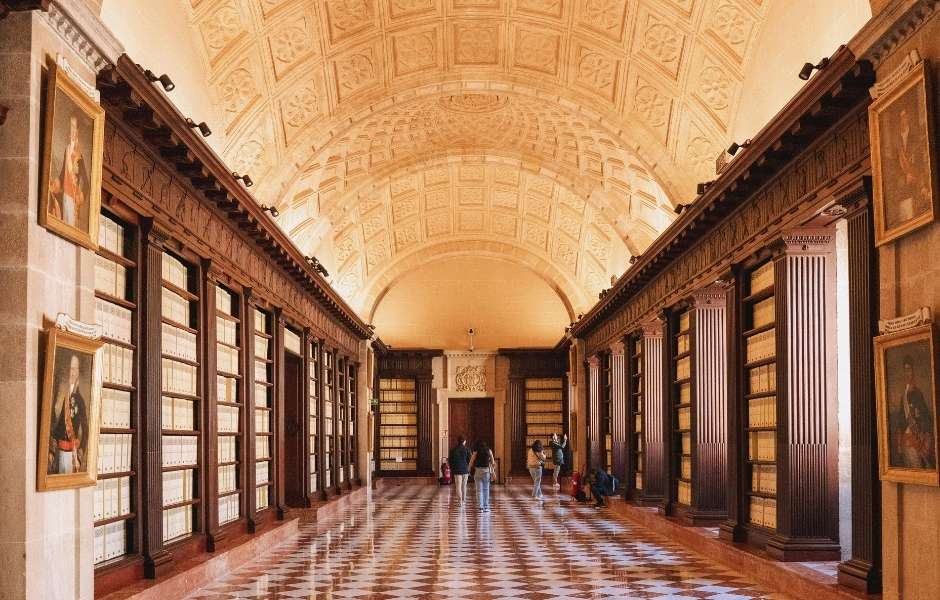 Visitors walking through a grand vaulted archive hall lined with floor-to-ceiling document storage, representing large-scale archival collections requiring structured management