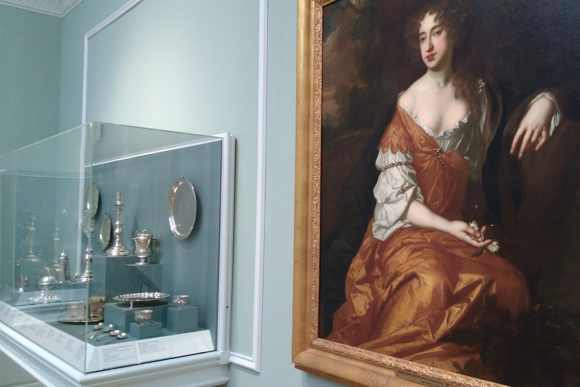 portrait of a woman next to her collection of silverware in a museum exhibition.