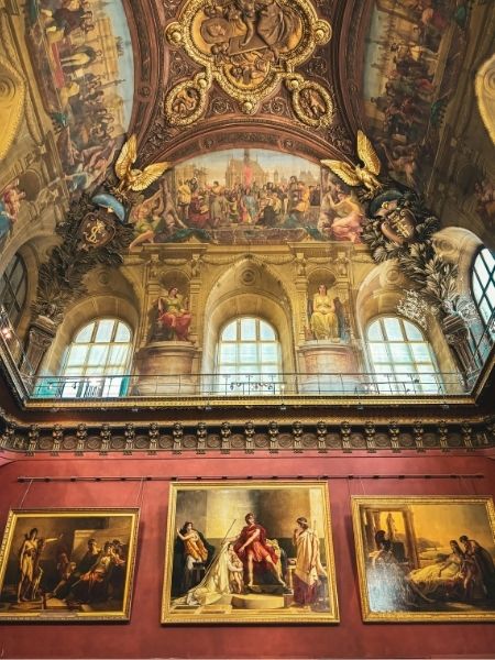 An ornate gallery interior with elaborately painted ceilings and large framed paintings displayed on deep red walls