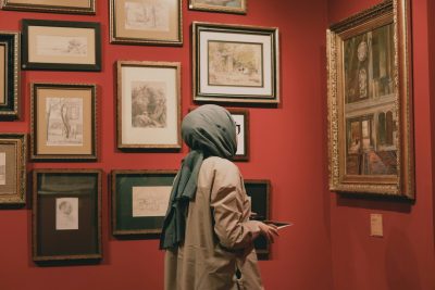 A visitor holding a tablet while viewing framed works in a red-walled gallery, representing public-facing access to collection media governed by rights and usage permissions