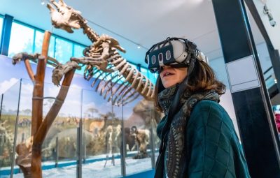 A visitor wearing a VR headset in a natural history exhibition space with a large fossil skeleton on display